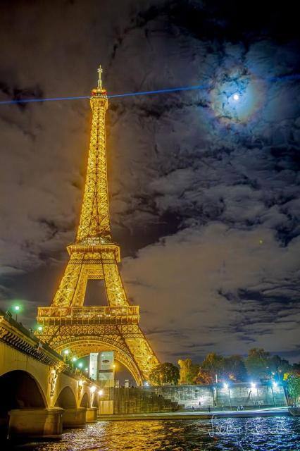 The famous Eiffel Tower in Paris, France is pictured on the left lit up in gold at night. A blue laser shines out from the top. Clouds dot the background sky. The Moon is also visible through the clouds, but is circled by colorful rings: a lunar corona.