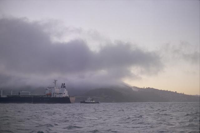 A large ship followed by a smaller vessel are leaving the frame. Behind them, green land covered in low hanging clouds, tinted purple by the setting sun. One distant star is already visible in the sky.