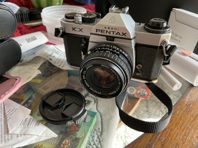 A Pentax KX 35mm camera placed on a glass table, with the lens cap removed, the lens is a SMC Pentax-M 1:1.7 50mm 