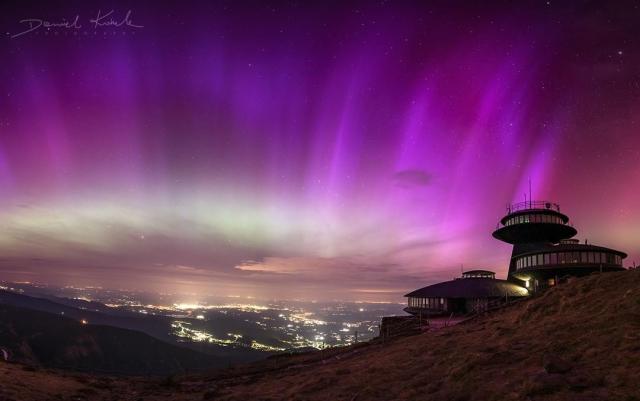 Purple striped aurora cover a star filled sky. Mountain peaks are visible on the sides, as well as a futuristic looking building on the right side. City lights are visible in the valley down below.