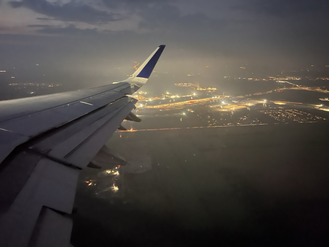 Night photograph from an airplane window, about 2000 feet in altitude, showing a large factory with several blazing white lights in a glowing mist.