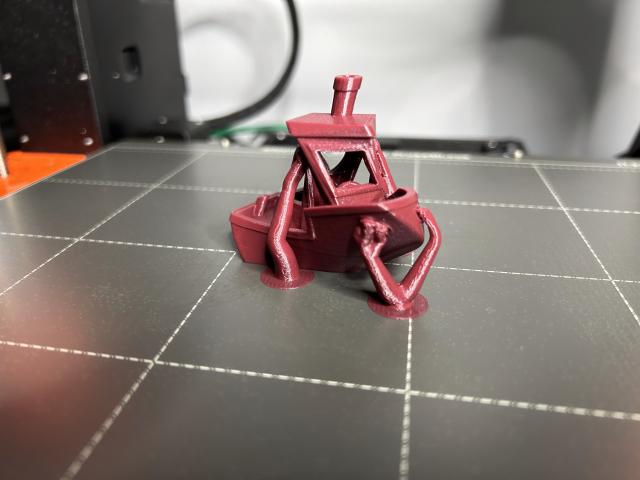 A merlot red PLA benchy on the printer bed with organic supports still attached.  In this photo, the bow is seen overhanging the starboard side of the hull , and we're looking in through the front window.