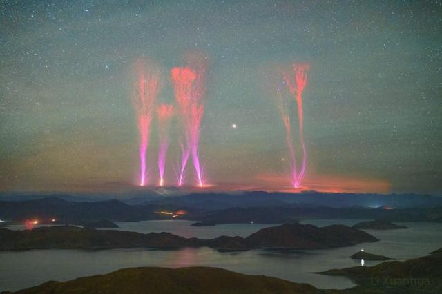 A landscape showing a night sky over distant mountains is shown. Lakes dot the foreground in front of the mountains. Extending from above the mountains into the night sky are six bright jets. The jets are violet at the bottom but red at the top.