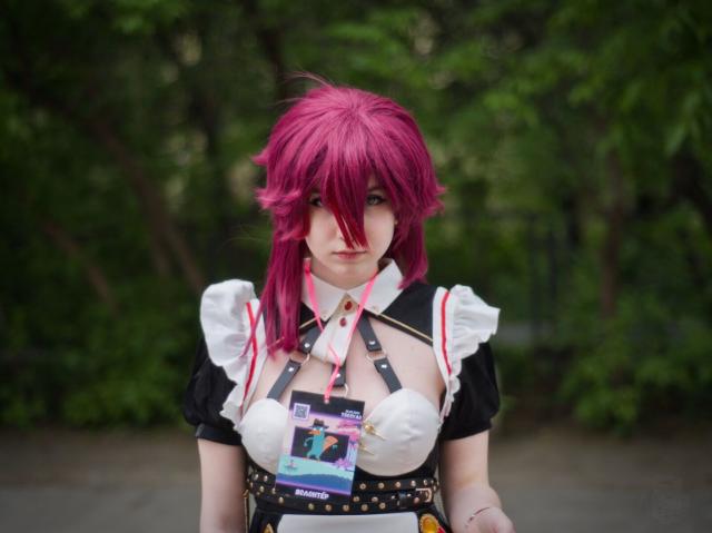 A waist-height portrait of a young woman cosplaying Rosaria from “Genshin Impact” game. She wears black dress with short sleeves, white apron, and black leather belt with rivets made of yellow metal. Her magenta hair is shoulder-length and one strand falls onto her nose. Her head tilted slightly forward. She does not smile and looks into the camera seriously.