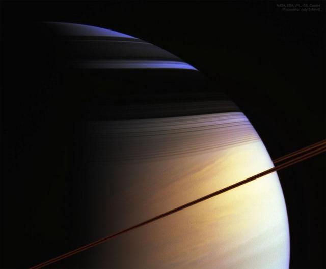 Saturn is shown taking up most of the frame. Most of the planet appears a banded gold. A thin line that is the rings appears dark brown and runs diagonally from the lower left. The upper part has dark bands which are shadows and behind the shadows the color of Saturn's atmosphere appears blue.