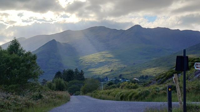 A small road leads up towards mountains. 2 beams of sunlight stream down from a sky of clouds with some blue, lighting up the slope of the valley in green. Majestic and otherworldly appearance. 