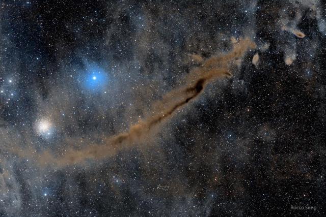 A busy starfield is shown which an elongated brown nebula running diagonally from the lower left to the upper right. A bright blue star and a star cluster appear above the nebula.