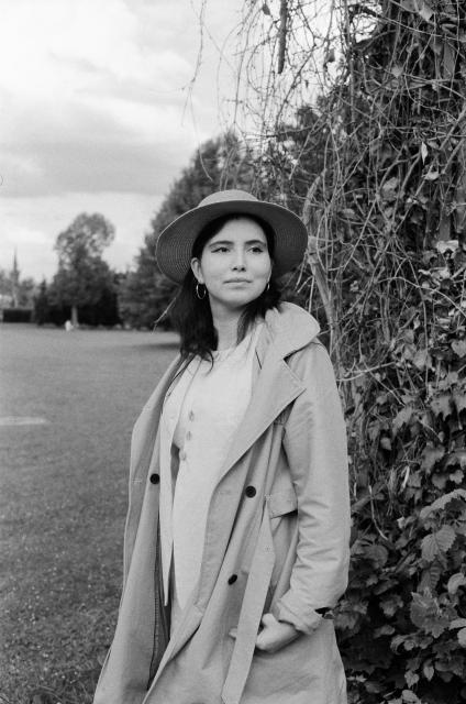 A black and white photo of a woman with black hair and hoop earrings, wearing a straw hat, a long trenchcoat and a waistcoat underneath.