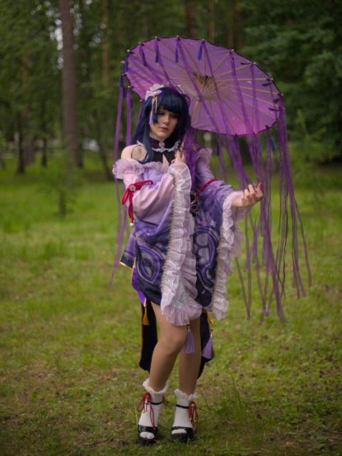 A portrait of a young woman cosplaying Raiden Makoto from “Genshin Impact”. She wears short lilac dress with long and very wide semi-transparent sleeves, white short socks and black sandals. She has chest-length blue hair and blue eyes. In the right hand she holds opened japanese-style lilac umbrella with long stripes of the same color hanging down from the umbrella and with blue tassels. She stretched out her left hand as if trying to feel rain outside the umbrella and holds some lilac stripes from umbrella in the palm of her hand. She looks at her left hand.