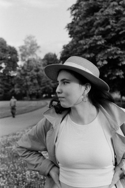 A black and white photo of a woman with black hair, hoop earrings, a straw hat, wearing a trenchcoat, and a tank top, looking to the side.