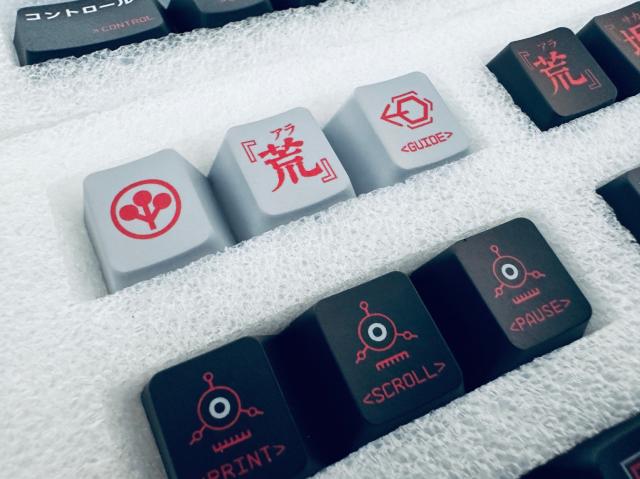 The image features a close-up view of several keyboard keycaps placed in a white foam tray. The keycaps are of different colors and feature unique and decorative designs.

From left to right:

1. A light gray keycap with a red symbol resembling stylized circles connected by lines.
2. A light gray keycap with red Japanese characters.
3. A light gray keycap with red and black geometric design, labeled "GUIDE" at the bottom.
4. A dark gray keycap with red Japanese characters, identical to the second key in design.
5. A dark gray keycap with a red outline around a small white circle, resembling an abstract eye or robot face, labeled "PRINT" at the bottom in red.
6. A dark gray keycap with a red outline around a small white circle similar to the previous key, labeled "SCROLL" at the bottom in red.
7. A dark gray keycap with a red outline around a small white circle similar to the previous keys, labeled "PAUSE" in red.

In the top left corner of the image, there is a partially visible dark gray keycap with Japanese characters and the word "> CONTROL" printed on it in red. 

The white foam tray has indentations customized to hold the keycaps securely in place.