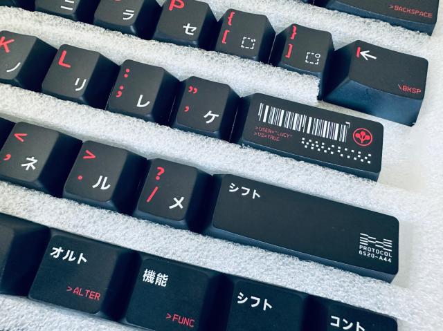 The image depicts a collection of black keyboard keycaps resting in a foam tray. The keycaps feature various legends in white and red text, symbols, and Japanese characters. Some keys contain unique designs, such as:

- A key with a barcode and the text ">USER='LUCY'", ">US=TRUE", and additional decorative symbols.
- A key labeled "ア" (Kana character) and another "コ" (Kana character).
- A key with a red dot and two angled symbols: "<" and ">"
- A long keycap with the text "シフト" (Shift in Japanese) and matching segments.
- Keys labeled with special terms such as "PROTOCOOL" followed by "6520-A44", "ALTER" in red below a long Japanese text, and "FUNC" in red beneath another Japanese text.

The keycaps are arranged in rows within the tray, offering an organized display of differing shapes and sizes appropriate for various keyboard functionalities.