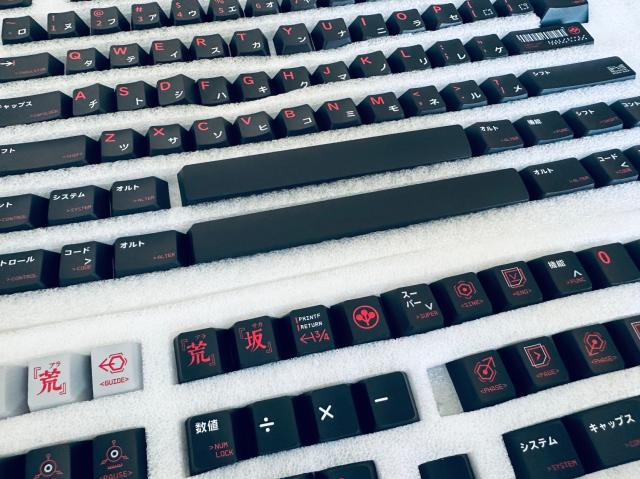 The image shows multiple black keycaps arranged in rows with red and white lettering, numbers, and symbols. Some keycaps have Japanese characters on them, while others have English letters. The keycaps are placed on a white foam surface for organization and protection. The rows include various typical keyboard keys such as alphabetic keys (A-Z), numeric keys, function keys, and modifier keys (Shift, Control, Alt). Some of the keys also have special characters or icons on them. The keycaps are intended for a keyboard, typically used for computers. The colors and symbols indicate this is a custom keycap set, likely for a mechanical keyboard enthusiast.