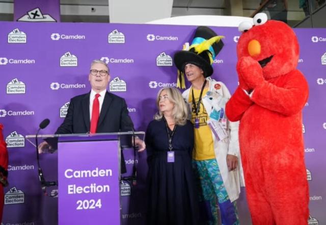 A photo of Keir Starmer winning his seat in parliament. He is pictured with a bloke in a huge, novelty hat and someone dressed as Elmo from Sesame Street. 