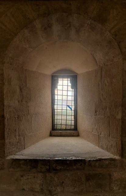 A narrow window set deep into a very thick wall. The panes are separated by a leaded grid, which is by no means orthogonally oriented. A blue strip of colour decorates one pane. The stone glows warmly with the incoming daylight.