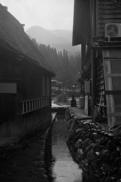Black and white film photo of a small canal between two houses in Shirakawago village with hills backlit by the sun in the background. 