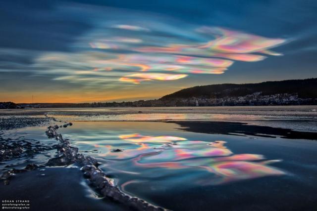 A landscape shows the sky above, shallow water in front, and hills to the right. The sunset sky is filled with beautifully multicolored clouds. These clouds are clearly reflected by the calm water in front.