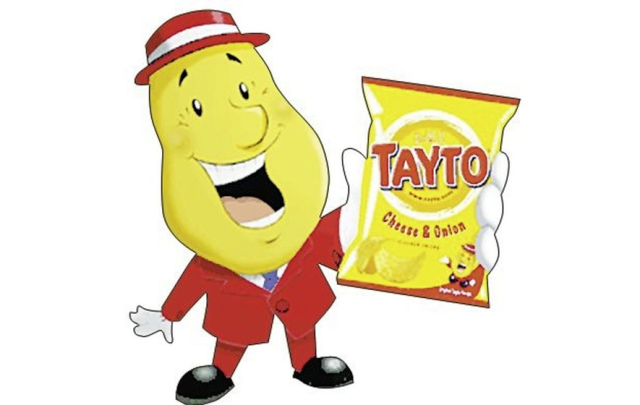 Northern Irish Mr Tayto wearing a red hat with a white stripe, a red jacket, white shirt, blue tie, red trousers, and black shoes. He's wearing white gloves and holding a bag of Tayto crisps