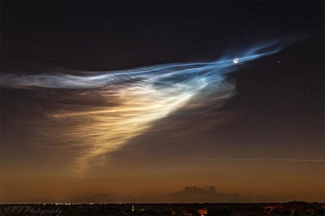 A dawn sky is shown that is black at the top and brown near the horizon. Wispy clouds converge on the right turning from a white to a blue hue. Near the apex of this convergence is a crescent moon.