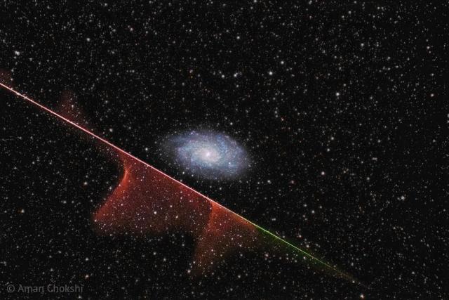 A distant spiral galaxy is seen in the image center. A multi-colored streak runs diagonally across the image from the upper left to the lower right. Parts of this streak have gas near it. The background is dark field filled with stars.