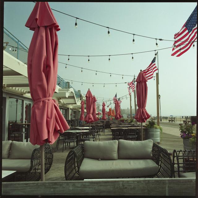 Couches and tables with chairs sit behind a barrier on the boardwalk. Umbrellas abound, all wrapped up and not deployed. A line of American flags punctuate the barrier.