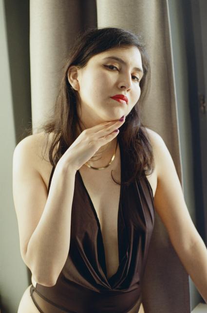 A beautiful woman with black hair, red lipstick, and purple nail polish, wearing a gold necklace and a black body, holding the back of her hand to her chin with a slightly tilted head.