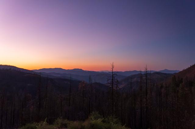The sun sets behind several layers of hills, amidst a semi-burned forest. [Fuji X-T5 / Tamron 18-300]