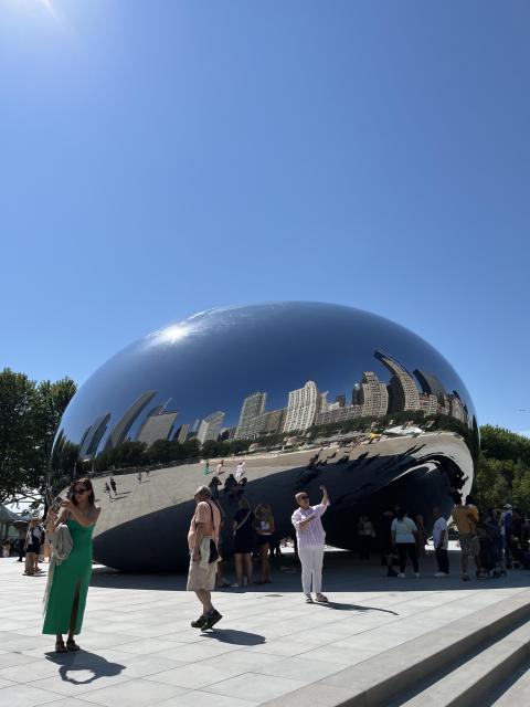 A photo of people taking selfies at The Bean at Millennium Park. The Bean is bean-shaped and has a mirrored surface that reflects everything all around it. In the photos reflection, you can see high rise buildings and lots of people.