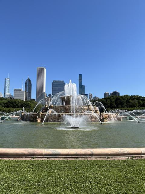 Spectacular Buckingham Fountain in Chicago, near the shores of Lake Michigan. The sky is a bright blue with no clouds, and you can see some high rise buildings in the background.