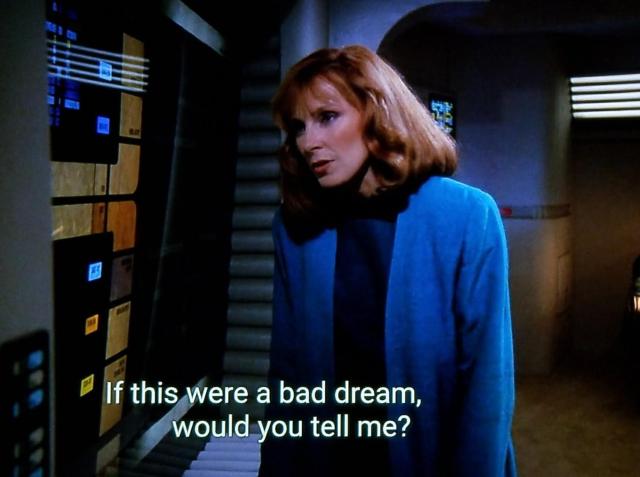 TNG scene. Candle enthusiast Dr. Beverly Crusher is standing in uniform alongside a wall computer panel leaning in as if she's speaking to it. The lights are dim. Closed caption reads, "If this were a bad dream, would you tell me?"