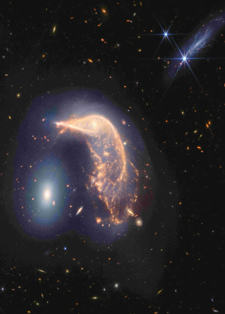 The distorted spiral galaxy at center, the Penguin, and the compact elliptical at left, the Egg, are locked in an active embrace. This near- and mid-infrared image combines data from NASA’s James Webb Space Telescope’s NIRCam (Near-Infrared Camera) and MIRI (Mid-Infrared Instrument), and marks the telescope’s second year of science. Webb’s view shows that their interaction is marked by a glow of scattered stars represented in blue. Known jointly as Arp 142, the galaxies made their first pass by one another between 25 and 75 million years ago, causing “fireworks,” or new star formation, in the Penguin. The galaxies are approximately the same mass, which is why one hasn’t consumed the other. NASA, ESA, CSA, STScI 