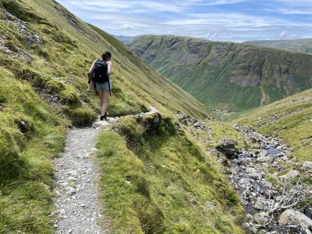 A steep trail on a green fell. In the background, more fells. Wife in front. 