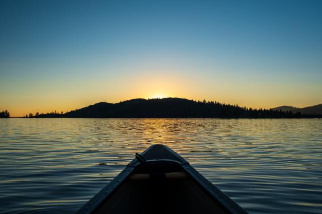 The bow of a canoe points across a lake at a hillside, behind which the sun is setting. [Fuji X-T5 / 16-55]