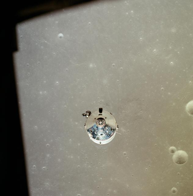 Color photo of the Apollo 11 Command and Service Module, taken from from the Landing Module, after undocking. The top of the module is a silver cone, seen nearly head on, attached to a white cylinder. The lunar surface – a yellowish gray, with craters – is in the background.
