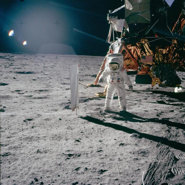 Color photo of the astronaut standing in front of the LM. The lunar surface appears gray and dusty. There is lens flare from the sun just out of the frame of the photo. The scene is reflected in the astronauts golden-colored visor.
