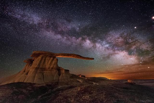 The band of the Milky Way runs across a night sky filled with stars. Colorful clouds are on the right horizon. A strange rock structure appears in the image center with a base and an extended arm that seems to point to the colorful horizon.