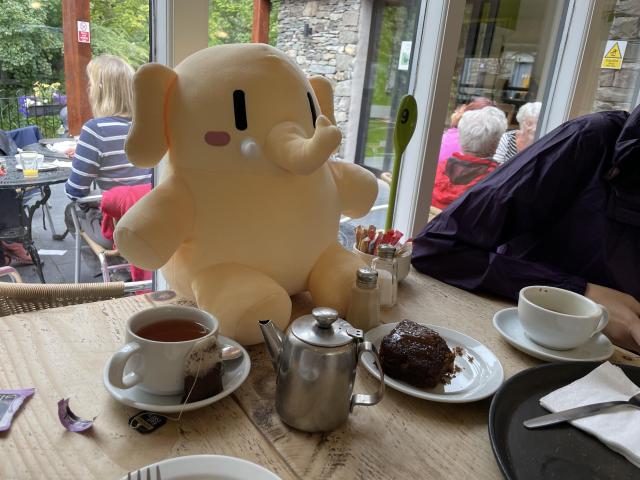 A stuffed Mastodon toy sitting on the table. In front of it, a cup of tea and cake on a plate. Behind him is a terrace with more tables. 