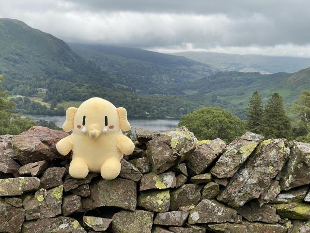 A stuffed Mastodon toy sitting on a stone wall overlooking a valley with a lake. 