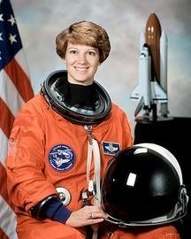 Astronaut Eileen Collins. First woman Space Shuttle Commander. Wearing her orange flight suit and holding her helmet. Model of the rocket with Shuttle and US flag can be seen behind her.
