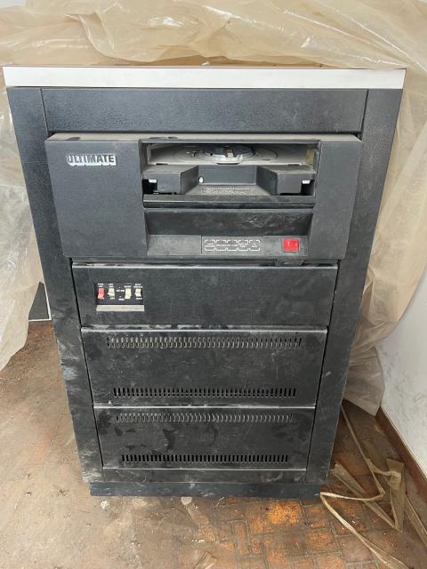 Photograph of the front of a half-rack-sized black computer sitting on the ground on rollers. In the top slot is a large tape reel reader, below that is the power control, and the modules below that are covered by a removable metal grille.