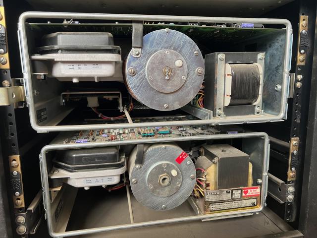 Front photograph of the two compute module trays with their protective metal grille removed. Each are identical; on the left is a large encapsulated hard drive. On the right appears to be a power supply. In the middle is a metal drum of unknown purpose. Both are topped by a large circuit board.