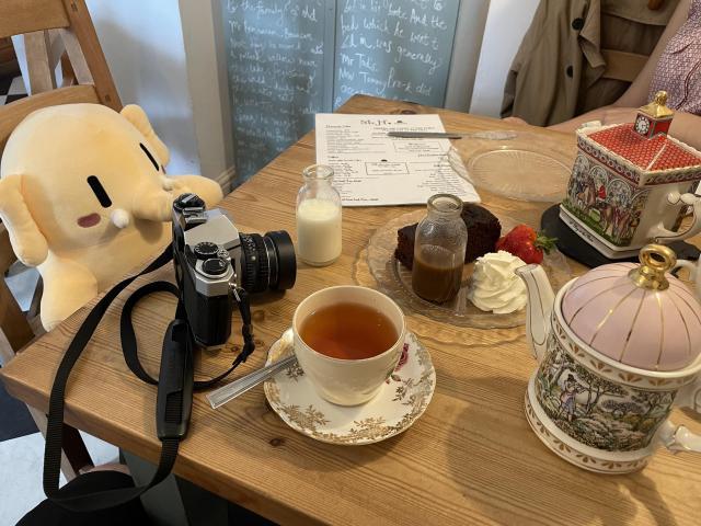 A stuffed Mastodon toy sitting behind a table with beautiful tea pots, a tea cup, sticky toffee pudding cake, and a menu where you can see the Mr. H logo. 