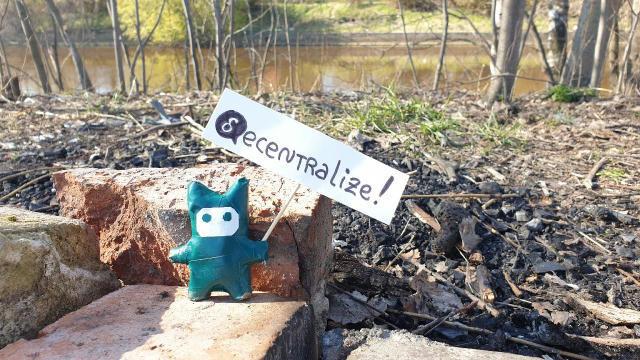 A small figure in a forest; it is holding a sign saying "decentralize!" with a delta chat logo.