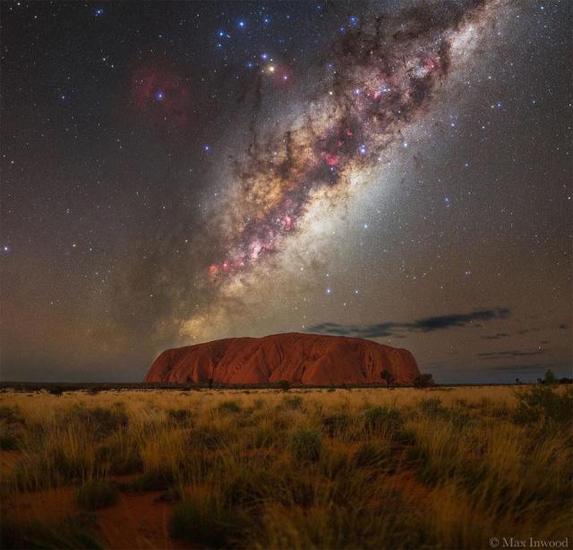 A starry sky is shown with the busy central band of our Milky Way Galaxy showing rising diagonally from the lower right. In the foreground are flat grasslands leading up to a huge orange rock mound named Uluru.