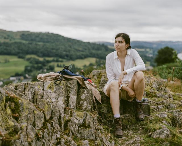 A woman with dark hair wearing shorts and a white long sleeve shirt sitting on top of a green rock. Behind her is a green valley and the edge of a body of water.