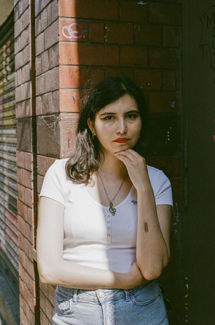 Beautiful woman wearing a white top, light blue jeans, a snake pendant, and red lipstick, standing in the shade of an entrance of a red brick building, with one hand holding her chin.