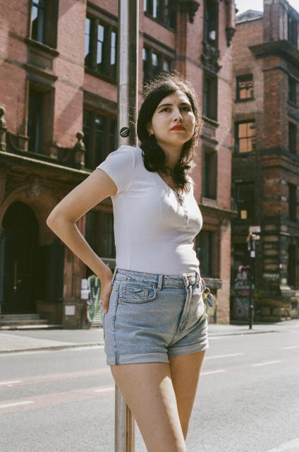 Beautiful woman wearing a white top, light blue jeans, a snake pendant, and red lipstick, leaning against a bus pole, with the sunlight illuminating her from behind.