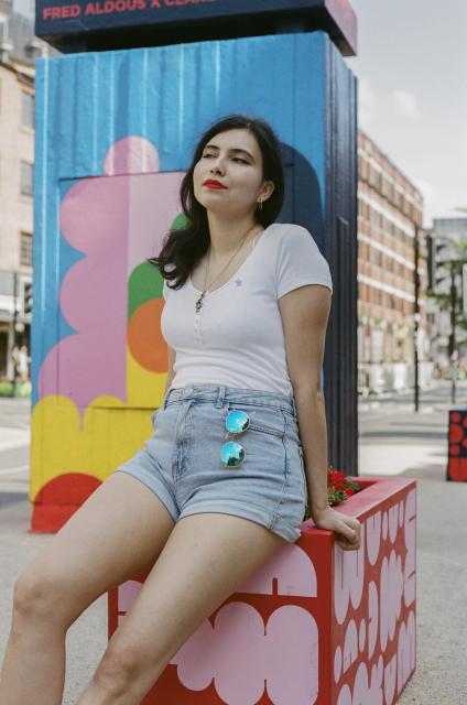 Beautiful woman wearing a white top, light blue jeans, a snake pendant, and red lipstick, sitting atop a red street planter, with a pair of blue tinted sunglasses tucked into her front pocket.