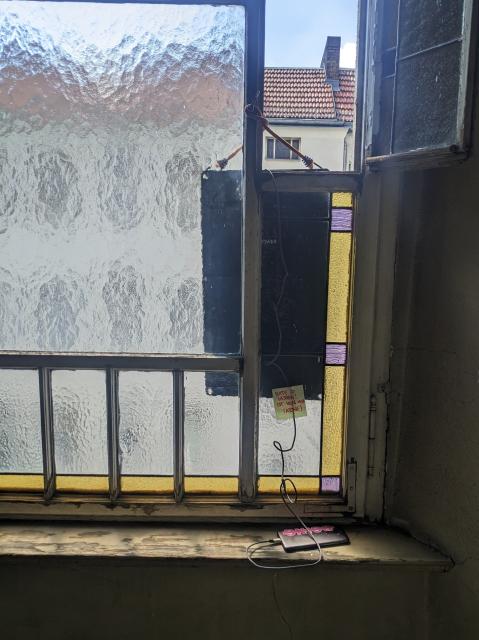 A smartphone is lying on a window sill. A part of the milky and colorful window behind it is open, and a solar charger is hanging outside. A small post-it note asks readers to please leave everything as it is.