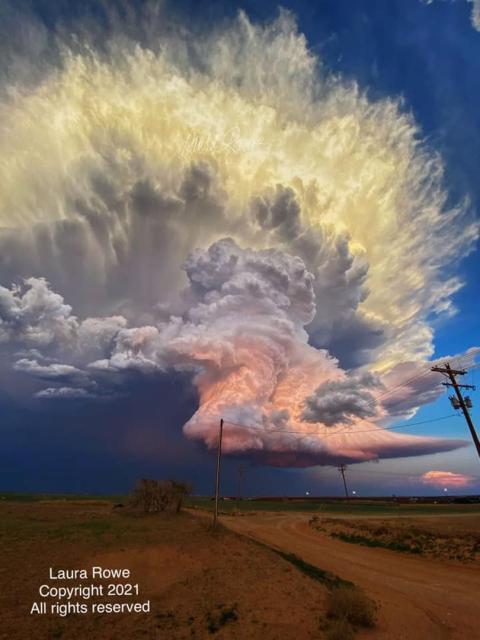 A large storm cloud is pictured hovering over a flat landscape with telephone poles. The background sky is blue. The cloud appears orange at the bottom, yellow at the top, white in the middle, with dark gray overtones all around.